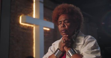 Afro-Amerikalı yaşlı kadın ellerini sımsıkı tutarak dua ederek gülümsüyor, kilisede yanan bir haçın önünde oturuyor.