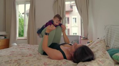 Anne yatakta uzanıyor, neşeli bebeği havaya kaldırıyor, el ele tutuşuyor, neşeli bir bağ kurma anı, aydınlık yatak odasında pencereli ve perdeli hassas bir aile bağı.