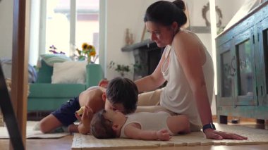 Anne yerde iki çocukla oturuyor, sıcak aile bağı, bebek mindere uzanıyor, büyük çocuk annesine yaslanıyor, şefkatli bir an, sevgi dolu bir bağ, rahat bir yuva.