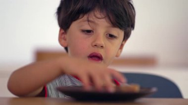 Atıştırmalık yiyen küçük çocuk, kurabiyelerden zevk alan, neşeli yüz ifadesi, evde atıştırma anı, yiyecek düşkünü çocuk, mutlu çocukluk anı, rahat atmosfer.