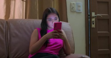 Kanepede oturan genç, akıllı telefonlu, sahne tavaları ekrandaki genç kıza bakıyor, teknoloji bağımlılığını ve çocuklara olan dijital bağımlılığı vurguluyor