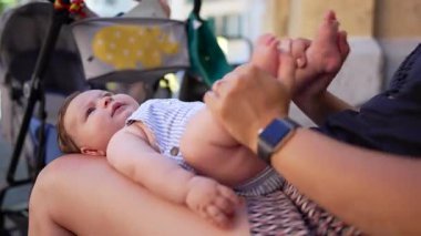 Anne kucağında bebeği tutuyor, gaz rahatsızlığını gidermek için bebek bacağını kaldırıyor, bebek bakıcılığı yapıyor, bebek bakıcılığı yapıyor, yakınlarda geziniyor.