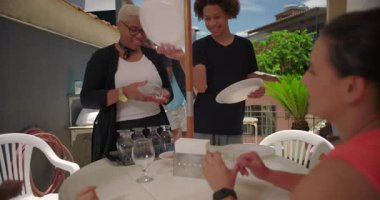 Bir grup arkadaş güneşli bir teras avlusunda tabak ve gözlüklü bir masa hazırlıyor, açık hava yemeği hazırlamak için birlikte çalışıyorlar.