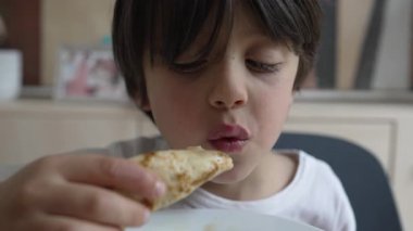 Genç çocuk lezzetli bir gözlemeden hoşlanıyor, ciddi bir ifadeyle lezzete odaklanıyor, evdeki değerli ve samimi bir yemek anını vurguluyor.