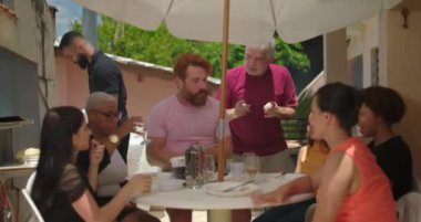 Farklı bir grup insan şemsiye altında açık hava yemeğinin tadını çıkarıyor sıcak ve misafirperver bir aile toplantısında yemek ve sohbet ediyorlar.