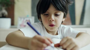 Mavi kalemle çizen çocuk, yaratıcılık sürecine odaklanmış, ev ortamında yakalanmış, çocukluk sanatsal aktivitelerinin neşe ve konsantrasyonunu vurguluyor