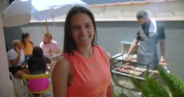 Arka bahçedeki barbeküde sıcacık gülümseyen bir kadın, aile ve arkadaşlar olarak neşe ve bağlantı sergiliyor, açık havada canlı bir kutlama için arka planda toplanıyor.