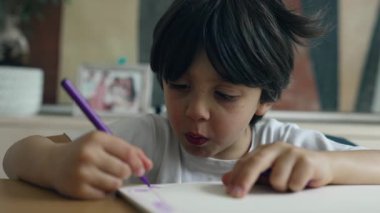 Çocuk mor bir kalemle çizime odaklanmış, yüzü ve eli görünürde, yaratıcılık anını rahat bir ev ortamında yakalama, konsantrasyon ve sanatsal ifadeyi vurgulama