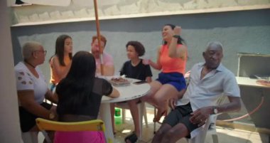 Barbekü ızgarası yemek pişirirken, farklı aile ve arkadaşlar arka planda oturup, dışarıda neşeli bir yaz kutlamasının tadını çıkarıyorlar.