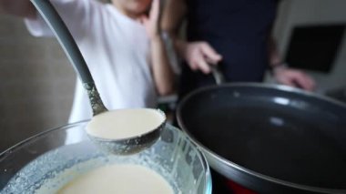 Çocuk krep hamurunu tavaya dökerken yakın plan, mutfak aktivitesi, işine odaklanmış çocuk, ebeveyn asistanı, beyaz tişört, sabah ışığı, ev ortamı, birlikte kahvaltı hazırlamak.