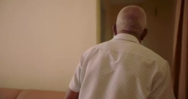 Yaşlı siyah bir adam koridorda yürüyor, arkadan bakıyor, desenli bir gömlek giyiyor, parlak ışıklı bir kapıdan giriyor.