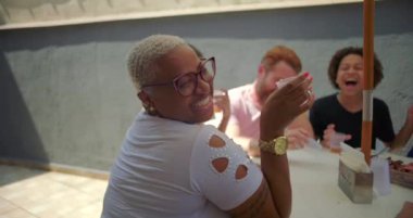 Gülümseyen Latin kökenli Afrikalı Latin bir kadın elinde bir kupayla yazın açık hava barbekü kutlamasında arkadaşlarıyla sohbet ederken başını çeviriyor.