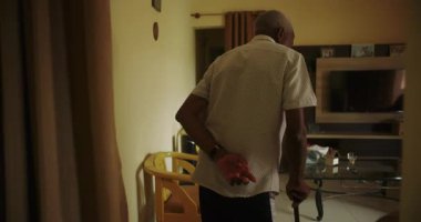 Oturma odasında elinde bastonla dolaşan yaşlı bir adam, dayanıklılık, bağımsızlık ve tanıdık bir ev ortamında yaşlanma zarafeti gösteriyor.