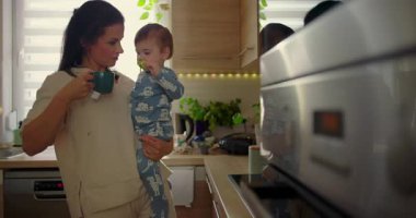 Anne mutfakta bebeği tutarken çocuk sıcak fincana uzanır, günlük aile rutini sırasında koruyucu ebeveynlik anı, sıcak bir evde bakım ve güvenlik