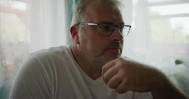 Adamın biri, çenesini elinde dinlendirirken, sakin ve rahat bir evde yumuşak yeşil perdeleri olan bir pencerenin yanında otururken düşünceli bir şekilde bakıyor.