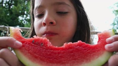 Önünde bir karpuz dilimi tutan genç bir kız bir ısırık daha almadan önce dokusunu inceliyor. Konsantre görünüşü bu açık havaya doğal ve samimi bir dokunuş katıyor.