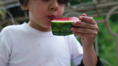 Genç çocuk hevesle bir dilim karpuz yiyor, sulu meyveyi ısırıyor. Odaklanmış ifadesi, bu ferahlatıcı atıştırmalığın doğal ortamdaki keyfini vurguluyor.