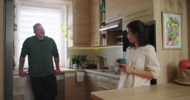 Kadın ve erkek, modern bir mutfakta rahat bir şekilde sohbet ediyorlar. Elinde kahve fincanı tutan kadınla birlikte. Parlak ve davetkar bir ortamda.