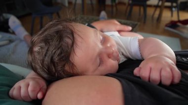 Bebek annesinin göğsünde mışıl mışıl uyuyor, kolları rahat, başı yana dönmüş, rahat bir aile ortamında hassas ve sessiz bir bağ kurma anını canlandırıyor.