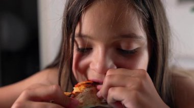 Bir parça pizza kabuğunu ısıran genç bir kız, iki eliyle tutuyor, zevk anında gözlerini kapatıyor, bir masada oturuyor, yüz ifadesine odaklanıyor.