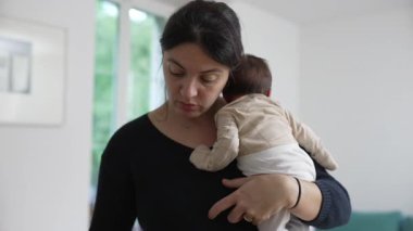 Anne beşikte, huzurlu bir ev ortamında sevgi ve bağlantı gösteriyor, anne bebeği nazikçe omzunda tutuyor.