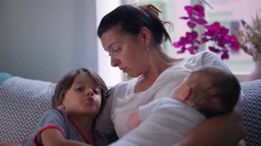 Anne, küçük oğluyla konuşurken bebeğini kucağına alıyor, birlikte kaliteli zaman geçiriyor ve sıcak bir ev ortamında samimi bir bağ kuruyor.