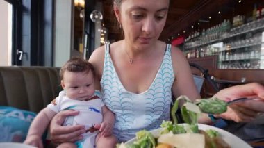 Bebek, restoranda salata yerken annesinin kucağında oturuyor. Sıradan bir yemek sırasında şefkatli ve şefkatli bir aile anı yaşıyor.