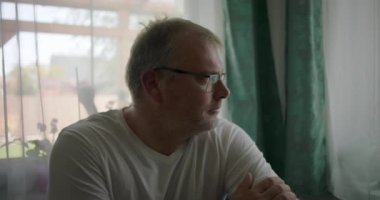 Pencere kenarında oturan bir adam, derin düşüncelere dalmış, elleri çapraz, sakin atmosfer kapalı, pencere ve perdeler yansıtıcı bir ruh halini çerçeveliyor.