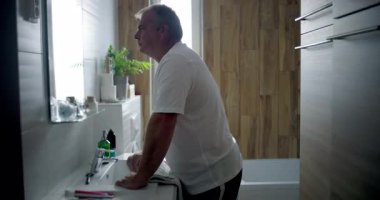 Profil görünümlü bir adam banyoda öne doğru eğiliyor, derin düşüncelere dalmış, içi beyaz fayanslarla dolu bir banyo ve ahşap kaplama, sessiz ve yansıtıcı kişisel bir an.