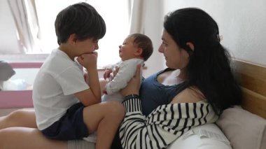 Küçük oğlu ve bebeği olan anne yatakta oturuyor, aile içi etkileşim, sevgi dolu bir an, rahat bir ev, kardeş bağı, anne bakımı, ev içi aile hayatı, duygusal bağ, hassas aşk