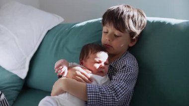 Küçük çocuk, yeni doğan kardeşini sevgiyle kanepede tutuyor ve sıcak bir yuva kuran kardeşler arasındaki hassas ve koruyucu bağı vurguluyor.