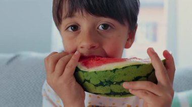 Küçük çocuk büyük bir karpuz dilimini ısırıyor, yakın çekim odağı, sulu yaz meyvesinin tadını çıkarıyor, sağlıklı beslenme, ferahlatıcı atıştırma, doğal ışık, iç mekan gündelik anı, çocukluk hazzı