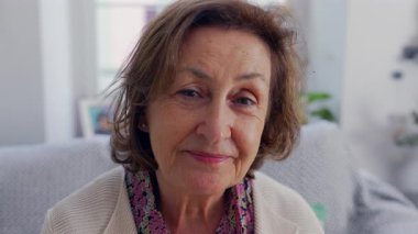 Yaşlı bir kadın nazikçe kameraya bakıyor, rahat bir evde oturuyor.