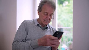 Pencerenin kenarında oturan yaşlı bir adam, akıllı telefonuna odaklanmış. Güneş ışığı içeri akarak, sakin, yeşil bir arka planda dalgın ifadesini vurgular.