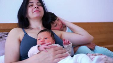Gülümseyen anne, iki küçük çocuğu yataktayken rahatlar, bebeğini kollarında tutarken büyük çocuk omzuna yaslanır, sevgi ve sıcaklıkla dolu dokunaklı bir aile anını gösterir.