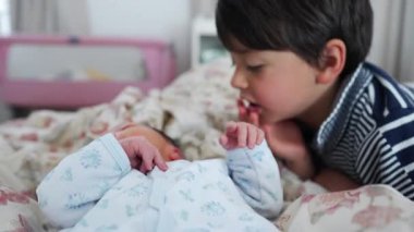 Ağabeyim yeni doğmuş bebekle yatakta nazikçe etkileşime geçiyor. Merakını ve şefkatini ifade ediyor. Yatak odası ortamını, kardeş bağlarını, sevgi dolu ve koruyucu jestleri, aile anlarını.