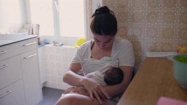 Anne yeni doğmuş bebeği mutfakta emziriyor, bir anlığına bakım ve bakım yapıyor. Yumuşak doğal ışıkla konforlu ev ortamında bağ ve sevgi dolu bir ortam vurgulanıyor