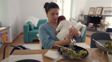 Anne yemek sırasında bebeği kucağında tutarken sıcacık ev ortamında bakım ve besinleri dengelemek, erken anne olmanın zorluklarını ve gerçeklerini göstermek, gündüz sahnesi