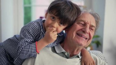 Dedeni arkadan kucaklayan çocuk, hem gülümsüyor hem de evdeki kanepede neşeli bir anın tadını çıkarıyor.