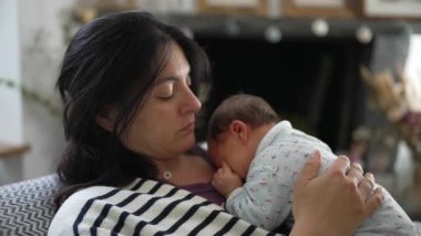 Anne göğsünde uyuyan yeni doğan bebeği tutuyor, huzurlu ve sevgi dolu bir anı paylaşıyor, sıcak bir aile ortamında güçlü bir bağ ve rahatlığı vurguluyor.
