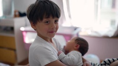 Ağabeyi gülümserken kucağında bebek kardeş, sıcak aile etkileşimi, sevgi dolu bir an, koruyucu bakım, ev ortamları, kardeş bağı, erken çocukluk, doğal ışık.