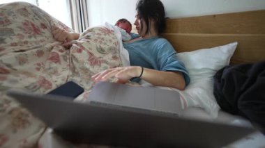 Anne göğsünde yeni doğmuş bir bebekle yatakta, dizüstü bilgisayara odaklanmış, rahat ve huzurlu bir şekilde ev ortamında birden fazla iş yapıyor, gösterişli bir bakım ve iş dengesi, samimi bir aile anı