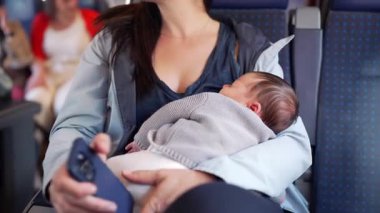 Annem trende seyahat ederken yeni doğmuş bebeği beşikte tutuyor. Bebek, yumuşak gri kazak giymiş, telefonunu tutarken sessizce kucağında uyuyor.