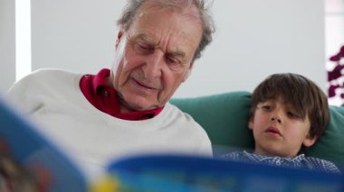 Büyükbabanın torununa resim kitabı okuduğu yakın plan çekimde, ikisi de hikâyeye odaklanmış, bir yuva ortamında nesiller arasında sevgi ve eğitim alışverişi sergiliyor..