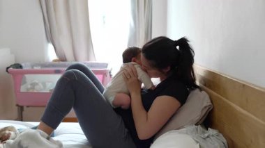 Anne bebeğini kucağında tutarken yatakta uzanıyor arka plandaki pencereden gün ışığıyla şefkatli bir öpücük paylaşıyor. Yumuşak ışık perdeleri ve pembe bebek beşiği olan rahat bir ev ortamı