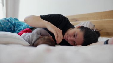Anne bir yatakta uzanıyor, bebek dinlenirken sevgi dolu bir şekilde bebeğinin başına dokunuyor, sıcak ve davetkar bir yuva ortamında sakin ve besleyici bir etkileşimi temsil ediyor.