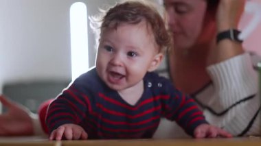 Bebek masada gülüyor ve emekliyor, heyecan ve enerji, anne arka planda gözlem yapıyor, neşeli etkileşim, neşeli bebek ev içi aile zamanlarında coşku dolu.