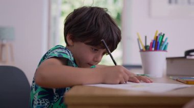Çocuk elinde kalemle sanat eserlerine odaklanmış, etrafı boya malzemeleriyle çevrili, evde yaratıcı aktivitelerle meşgul, yakındaki bir pencereden gelen doğal ışık.