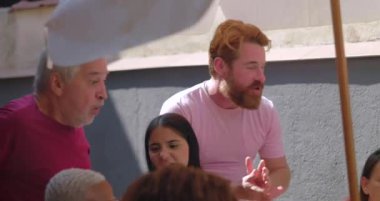 Çeşitli arkadaşlar canlı bir yemek sırasında dışarıda alkışlıyor ve gülüyor, bir şemsiye altında yapılan çok kültürlü bir toplantıda bağlantıyı, kutlamayı ve eğlenceyi vurguluyorlar.