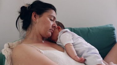 Anne ve bebek kanepede birlikte dinleniyor, başları nazikçe birbirine dokunuyor, huzurlu bir evde derin bir bağı, sıcaklığı ve huzuru simgeliyor.